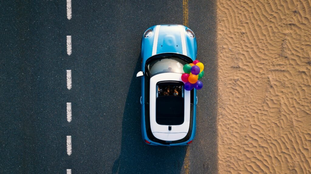 A Mini Cooper with colorful balloons drives along a desert road. Captured from above.
