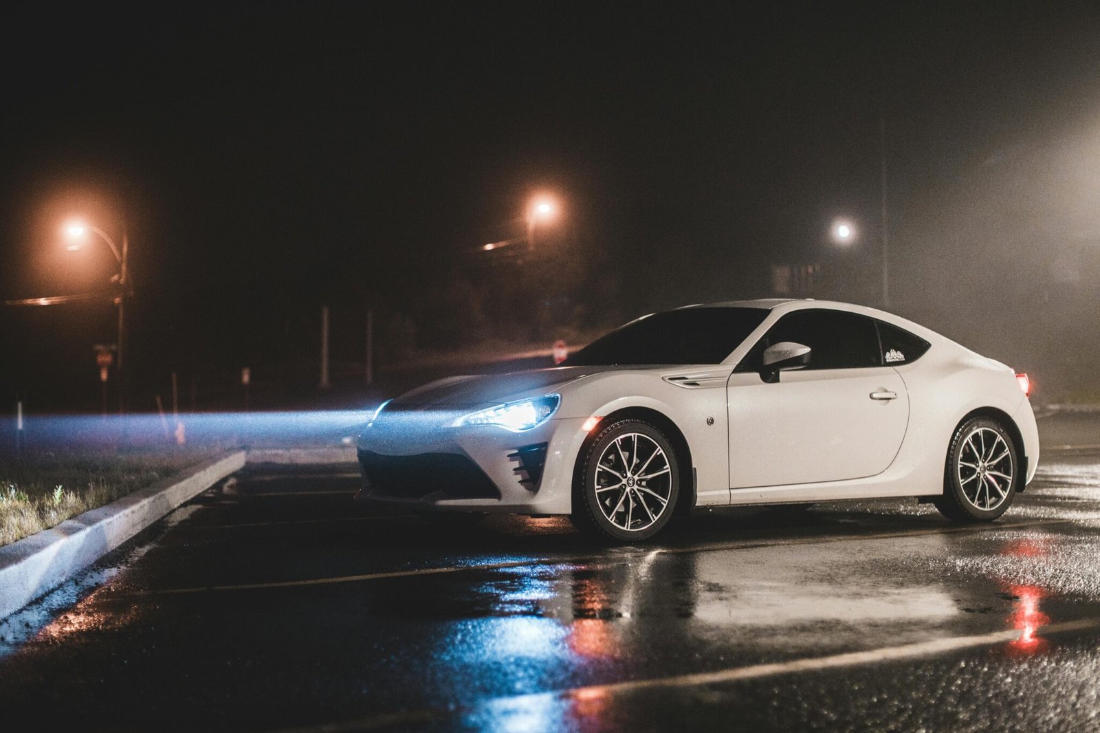 A modern white sports car parked on a wet street at night with glowing headlights.